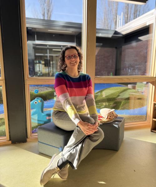 Image of woman with red glasses, brown hair, and striped sweater seated in a preschool classroom